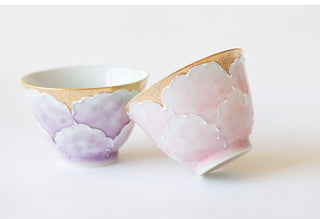 Two ceramic tea cups with scalloped floral designs, one in lavender purple and the other in soft pink, both featuring gold-gilded rims and interiors, set against a clean white background.