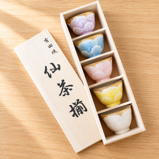 A set of five hand-painted ceramic tea cups in pastel shades of purple, blue, pink, yellow, and white, presented in a light wood box next to its lid on a wooden tabletop.