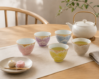 A set of five ceramic tea cups in pastel colors with gold accents, arranged on a beige runner on a wooden table next to a white teapot, with a small plate of sweets in the foreground.