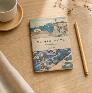 A hardcover notebook titled 'RO-BIKI NOTE Museum Series' with a Japanese ukiyo-e style illustration on the cover, styled on a light wood table next to a ceramic cup, a pencil, and a beige cloth.