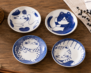 Four small ceramic dishes with cat designs on a wooden table with chopsticks and a tea cup.