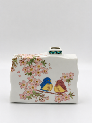 A white ceramic teapot with a lid, decorated with pink cherry blossoms and two small birds in blue, yellow, and brown, set against a plain light gray background.